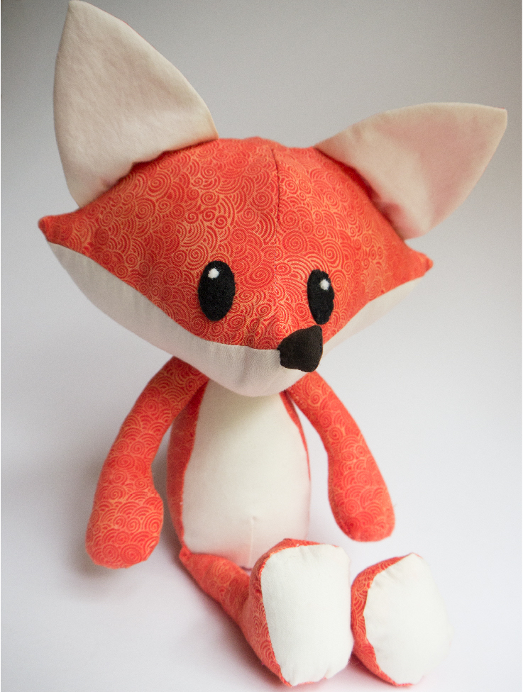 A photo of a stuffed fox with a pointy hand and large flat ears. It is made of patterned orange fabric with a white stomach, bottom of its paws, and ears.